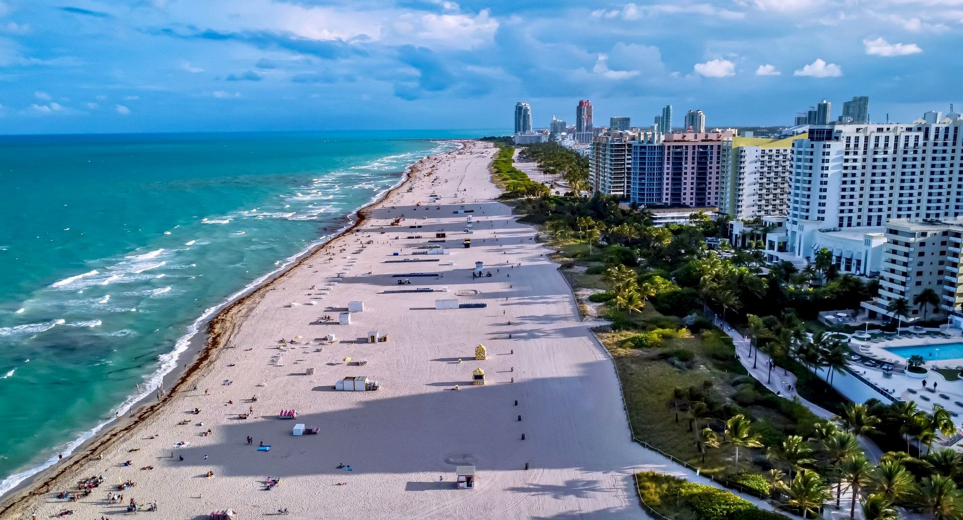 Aerial view of a sunny coastline with a sandy beach, ocean waves, palm trees, and high-rise buildings along the shore.
