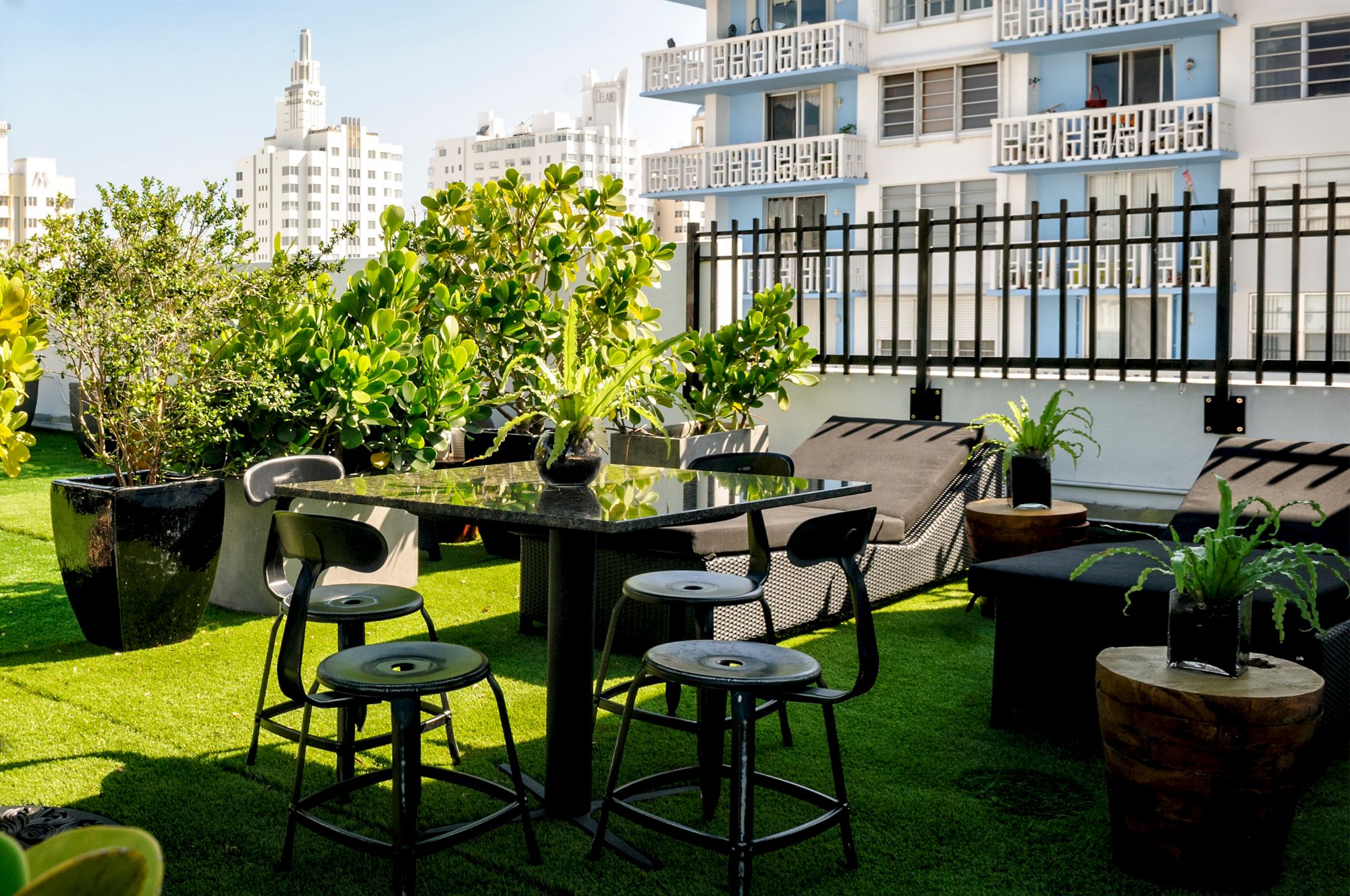 A sunny apartment balcony patio with a glass table, stools, potted greenery, and tall buildings in the background.