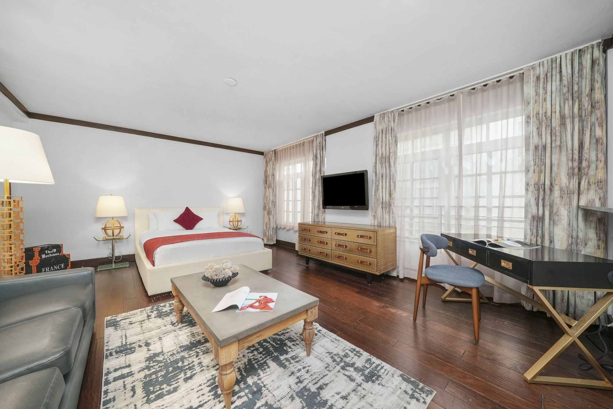 A modern hotel bedroom with a bed, lamps, sheer curtains, a wall TV, dresser, desk, chairs, and a coffee table.