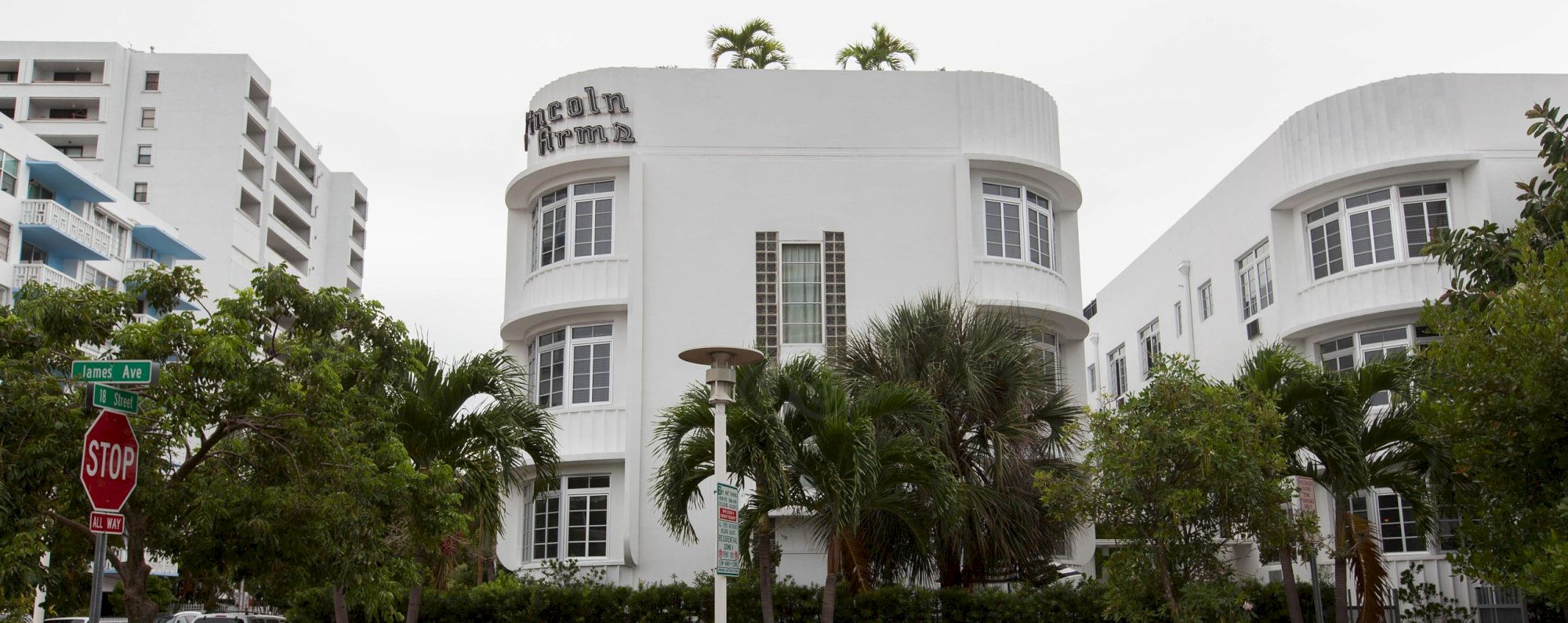 A white modern hotel building with &ldquo;Iconic Arr&hellip;&rdquo; on the facade, surrounded by palm trees, a red car, and a STOP sign in front.