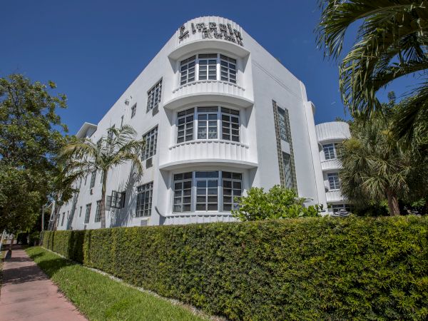 A modern white building with curved balconies and a rooftop sign, surrounded by lush palm trees under a blue sky.