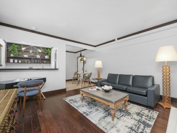 A modern living room with a gray sofa, coffee table, patterned rug, warm lamps, and an open kitchen shelf display.