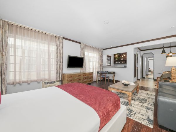 A bright hotel room with a bed, red blanket, TV on the wall, sheer curtains, and a living area with sofa, lamp, and coffee table.