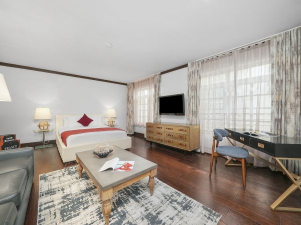 A bright hotel room with a bed, curtains, wall TV, chest dresser, desk, lamp, sofa, and a patterned rug.