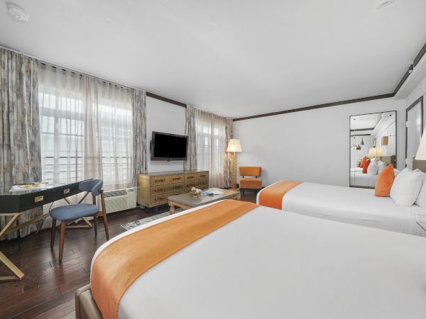 A modern hotel room with two beds, orange accents, curtains, a desk with chair, a TV, and a mirror reflecting the space.