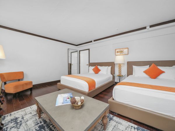 A bright hotel room with two beds, orange pillows and blankets, a glass side table, and warm lamps along the walls.