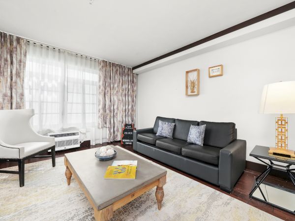 A bright living room with a black leather sofa, coffee table, patterned curtains, framed art, and a floor lamp.