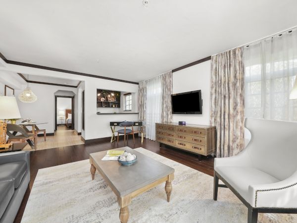 Bright modern living room with a wall-mounted TV, vintage-style console, coffee table, leather chairs, and sheer curtains.