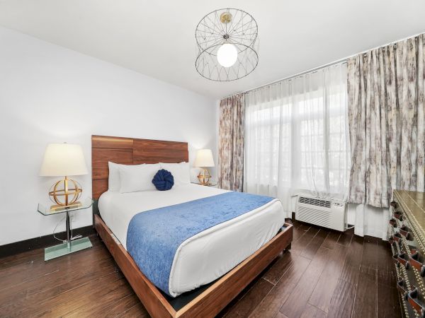 A modern bedroom with a wooden bed, blue blanket, globe pillows, bedside lamps, patterned curtains, and a ceiling fan light.