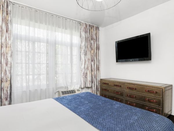 A bedroom with a blue bedspread, patterned curtains, a wall-mounted flat-screen TV, and a studded metal dresser.