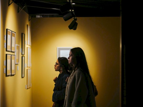 Two women in coats stand in a hallway under studio lights, facing framed displays on the wall.