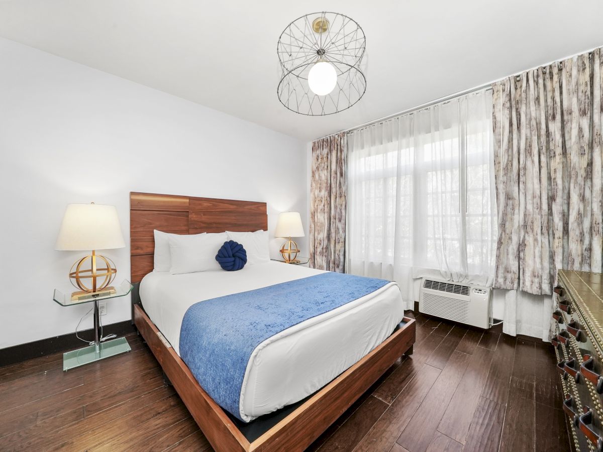 A bright bedroom with a wooden bed, blue throw, globe decor, pendant light, patterned curtains, and an AC unit by the window.