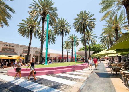 Sunny outdoor plaza with tall palm trees, turquoise light poles, umbrellas, and people walking and dining.