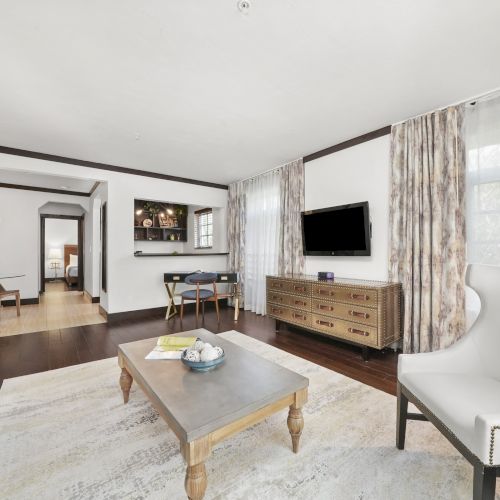 A modern living room with sofas, a coffee table, a wall-mounted TV, sheer curtains, and a studded media cabinet.