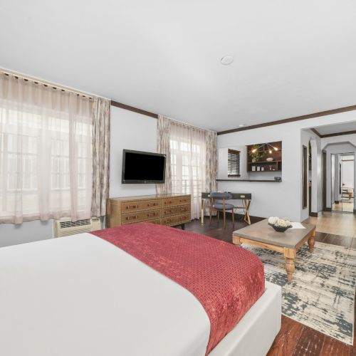 A bright bedroom with a red bedspread, curtains, wall TV, and an open view to a cozy living room with sofa and lights.
