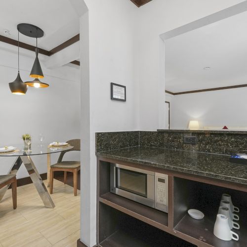A modern dining area and kitchen, with table settings, pendant lights, granite counter, oven, and small plants, in a bright room.