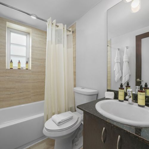A bright bathroom with a bathtub, white shower curtain, toilet, sink, and bottles on the counter beneath a mirror.