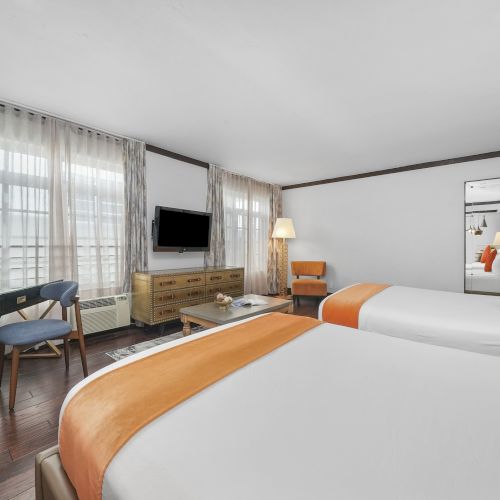 A modern hotel room with a large bed, orange pillows, desk, TV, and sheer curtains around windows.