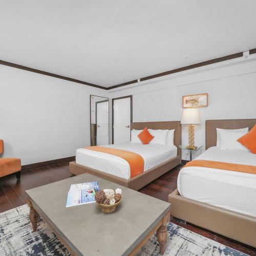 A bright hotel-style bedroom with two twin beds, orange accent pillows, lamps, and a glass-door closet, plus a coffee table.