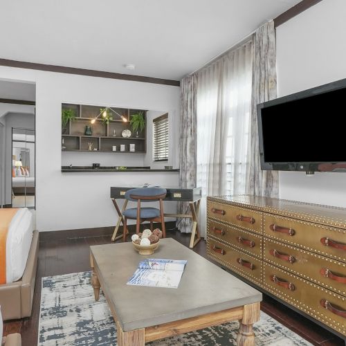 Bright living room with an orange sofa, coffee table, large wall TV, gold studded dresser, and window curtains.