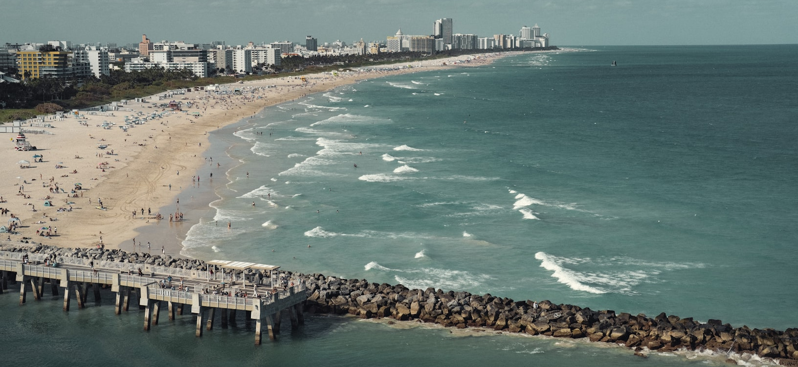 A wide sandy beach with crowds, gentle waves, and a rocky breakwater with a pier, facing a distant coastal city skyline.