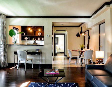 Modern living room with sofa, coffee table, and open dining area; built-in bar shelving and pendant lights brighten the space.