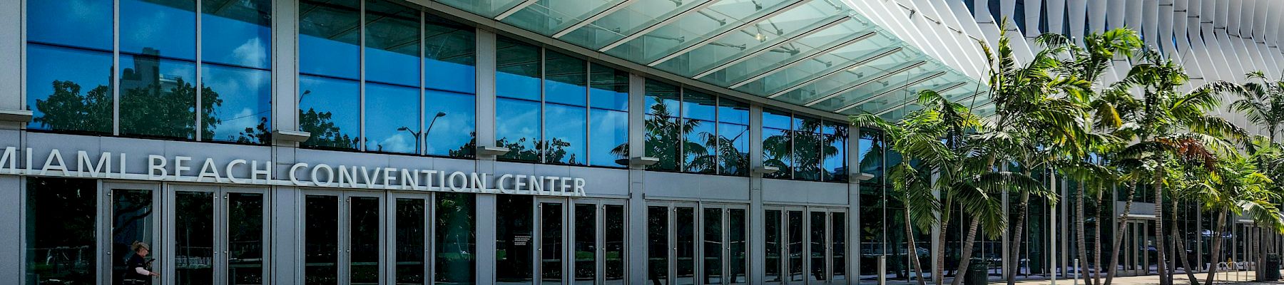 Miami Beach Convention Center entrance with glass awnings, tall white columns, and palm trees under a blue sky.