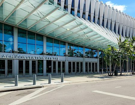 Miami Beach Convention Center entrance with glass awnings, tall white columns, and palm trees under a blue sky.