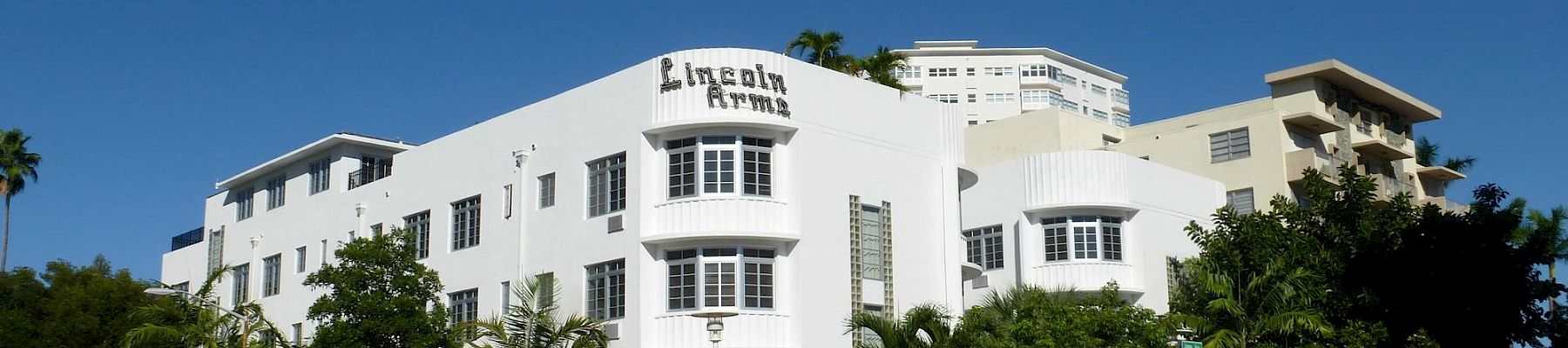 A sunny street view of the Lincoln Arms building, a white hotel with palm trees and parked cars under a clear blue sky.