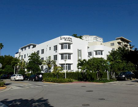 A sunny street view of the Lincoln Arms building, a white hotel with palm trees and parked cars under a clear blue sky.