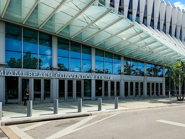 Exterior of the Miami Beach Convention Center with a glass facade, modern white fins, and palm trees under a blue sky.