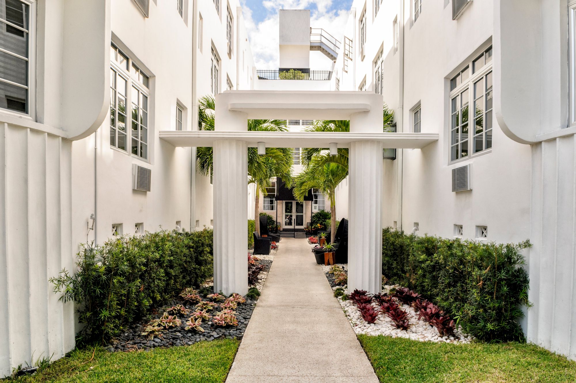 A bright modern courtyard walkway with white columns, lush tropical plants, and windows in surrounding buildings.
