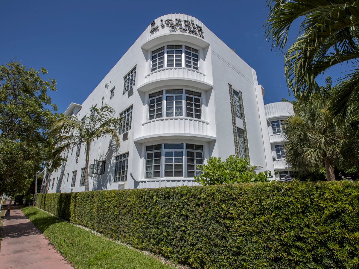 A modern white building with &ldquo;LE PLAZA&rdquo; signage on the rooftop, surrounded by palm trees and trimmed hedges under a blue sky.