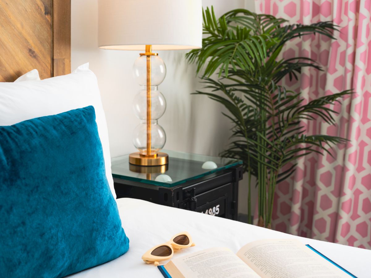 Cozy bedroom scene with a bed, blue pillow, tall lamp, potted palm, and pink geometric curtains.