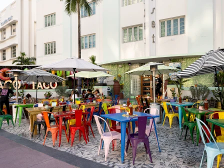An outdoor taco/caf&eacute; patio with colorful plastic chairs and tables under striped umbrellas beside a white building, in a sunny courtyard.