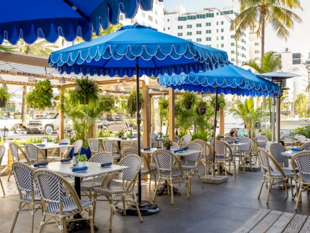 An outdoor patio restaurant with blue striped umbrellas, patterned metal chairs, palm trees, and buildings in the background.