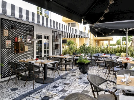 An outdoor patio dining area with round tables, black chairs, striped awnings, and lush potted plants lining the walkway.