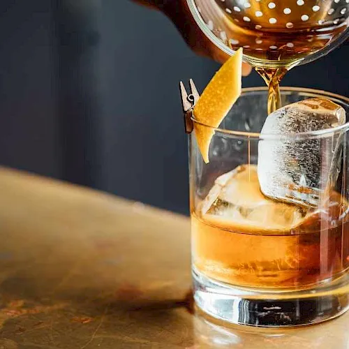 An amber cocktail is being poured over ice in a rocks glass, garnished with a citrus wedge for a bar drink finish.