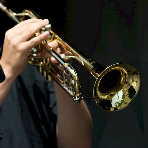 A musician plays a shiny brass trumpet, holding it up while sounding notes under stage lighting.