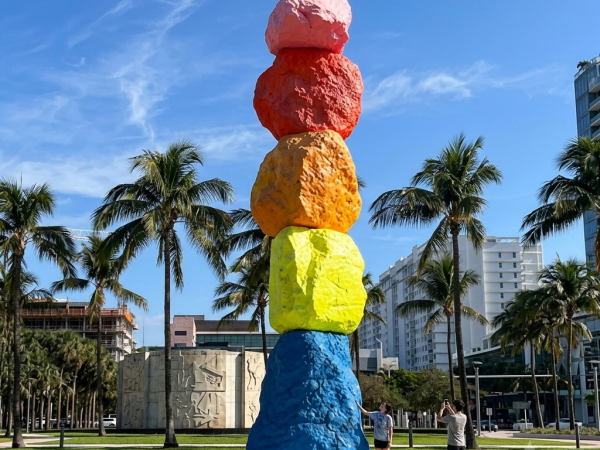A colorful stacked-stone sculpture towers in a sunny resort garden with palm trees, white buildings, and a couple walking nearby.