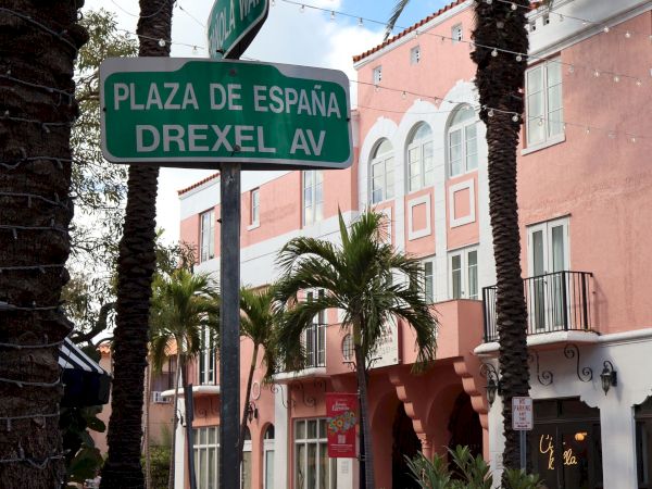 A street sign reading &ldquo;PLAZA DE ESPA&Ntilde;A DREXEL AV&rdquo; stands among palm trees and a sunny courtyard buildings backdrop.