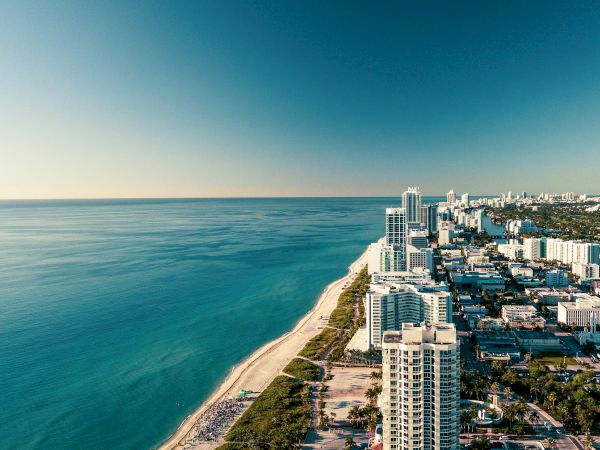 Aerial view of a sunny ocean coastline with sandy beach and high-rise buildings along the shore.