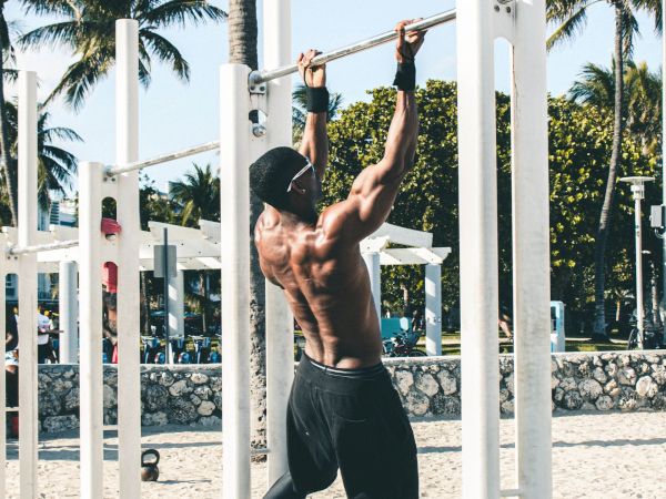 A shirtless man does pull-ups on an outdoor beach fitness bar set, with palm trees under a sunny sky.