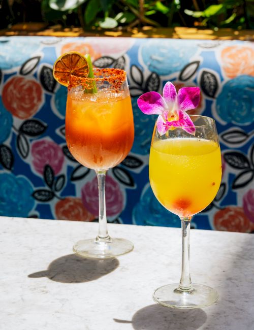 Two colorful tropical cocktails in wine glasses, garnished with citrus slices and an orchid flower, served on a sunny patio.