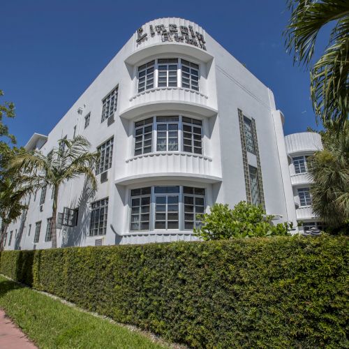 A modern white building with glass balconies and a rooftop sign, surrounded by manicured hedges and palm trees under a clear blue sky.
