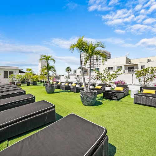 A sunny courtyard with green lawn, black lounge chairs with yellow cushions, palm trees, and white apartment buildings under blue sky.