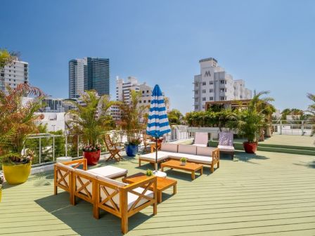 A rooftop terrace with wooden seating, planters, blue-striped umbrella, and palm trees overlooking a modern city skyline.
