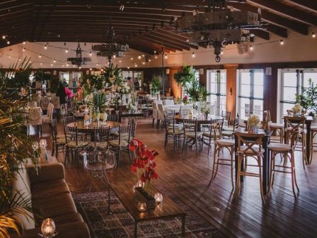A warmly lit event hall with neatly set tables, chairs, and lots of potted greenery and flowers for a reception.