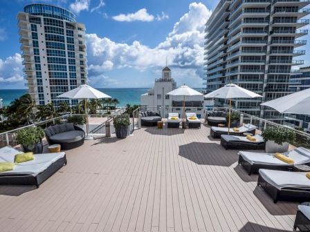 A sunny hotel rooftop terrace with lounge chairs and white umbrellas, facing the ocean between tall apartment towers.