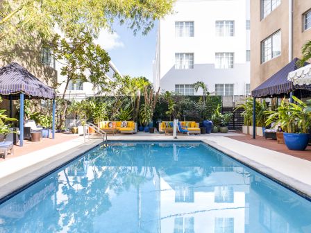 Sunny courtyard pool with blue water, lounge chairs under striped awnings, and lush tropical plants beside a white apartment building.